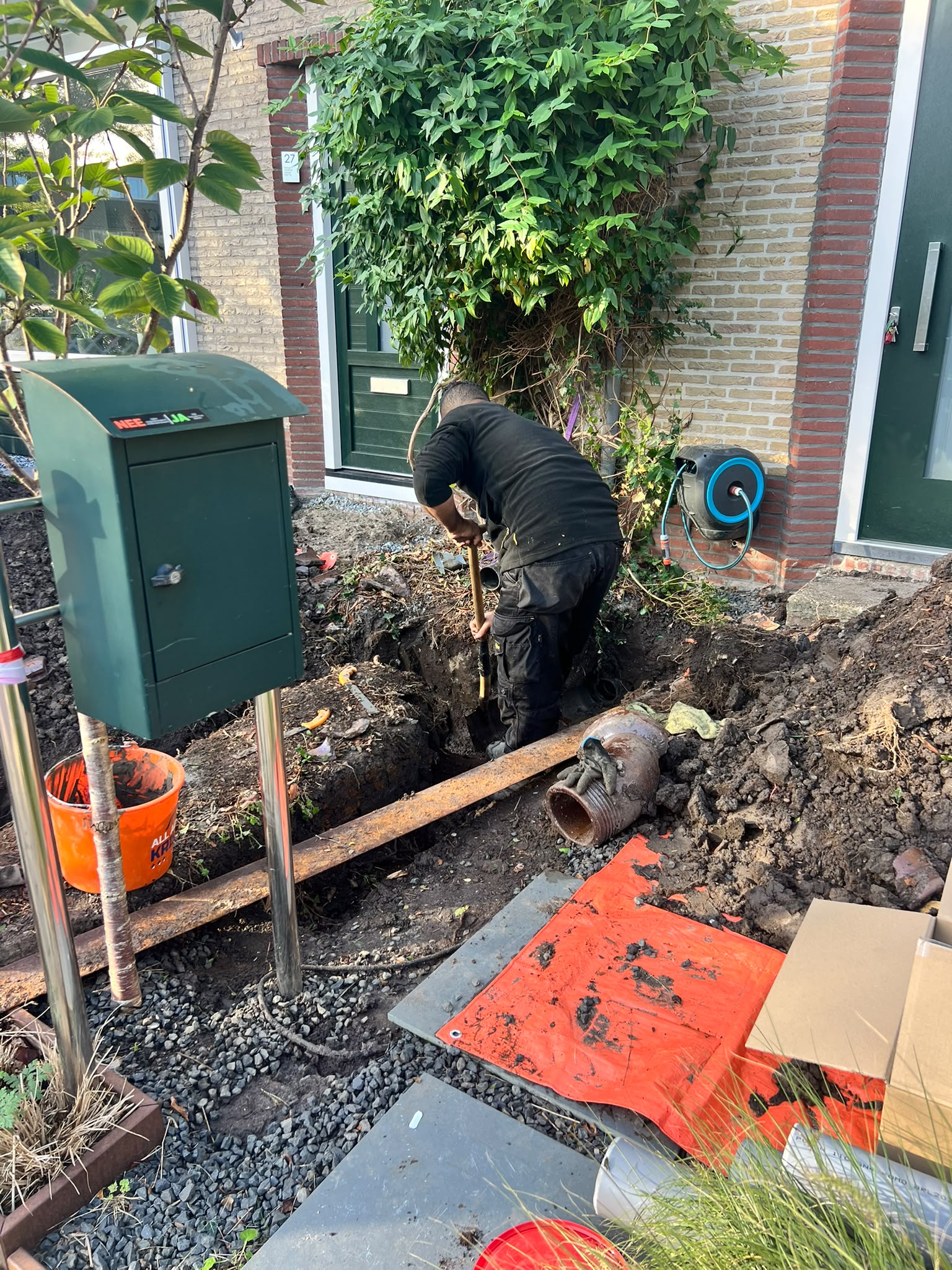 Monteur graaft een sleuf in de voortuin naast de brievenbus; oude rioolbuisdelen liggen ernaast