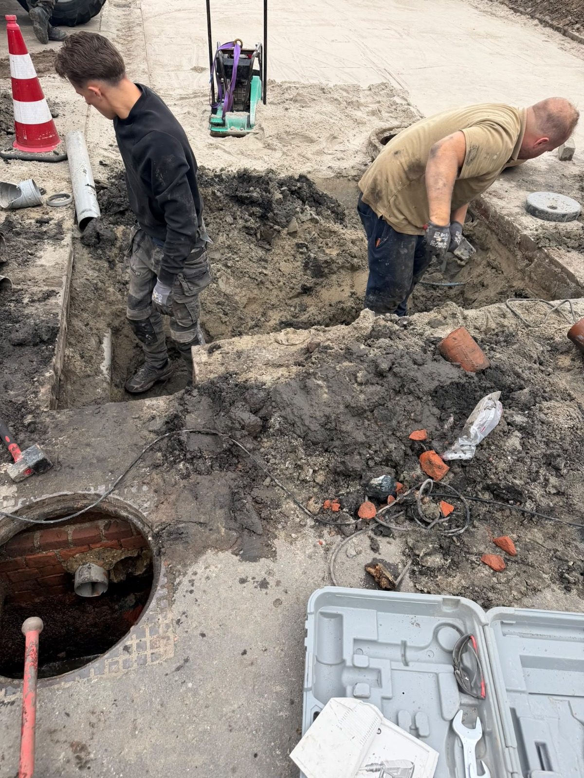Twee monteurs aan het werk bij opengebroken bestrating naast een open gemetselde rioolput